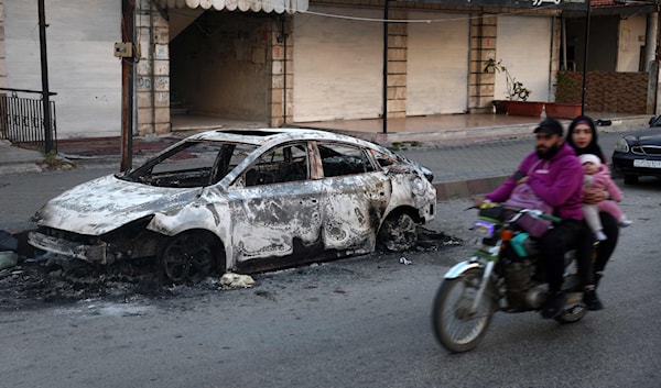 أفراد عائلة يركبون دراجة نارية يمرون بجانب سيارة متفحمة في بلدة جبلة في محافظة اللاذقية (أ ف ب)