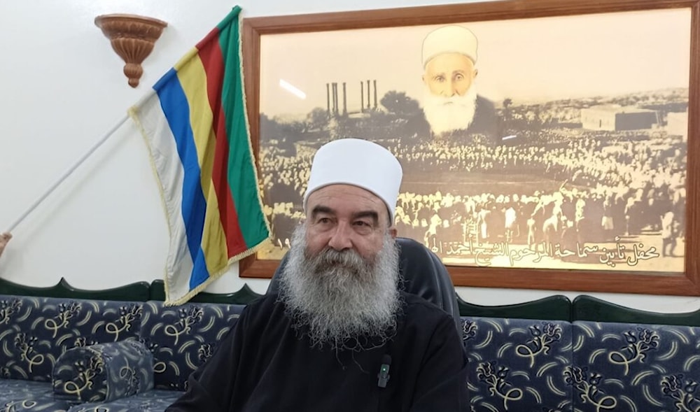 حكمت الهجري ويظهر خلفه صورة والده الشيخ أحمد الهجري الأول (من الويب)