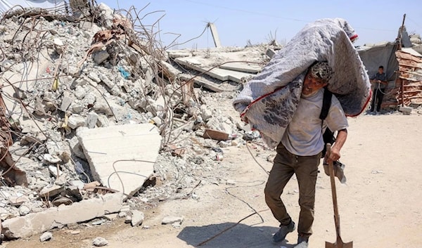 فلسطيني يحمل أمتعة على كتفه، بمحاذاة الأنقاض في مخيم للنازحين بغزة (أ ف ب)