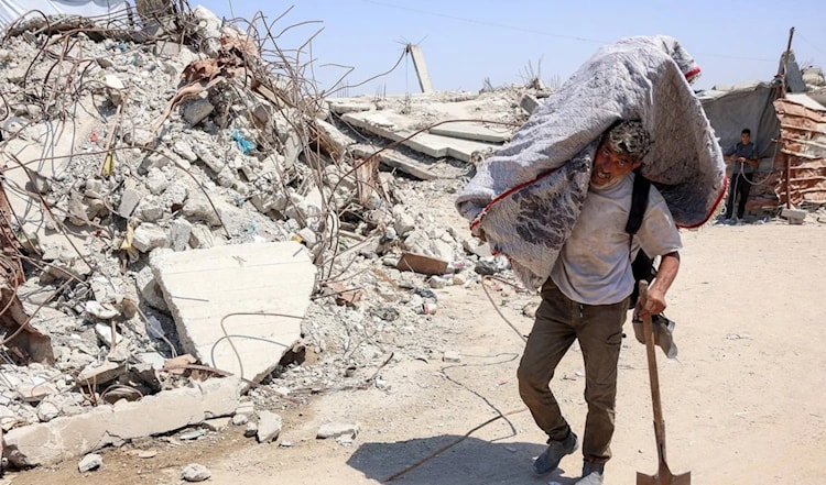 فلسطيني يحمل أمتعة على كتفه، بمحاذاة الأنقاض في مخيم للنازحين بغزة (أ ف ب)